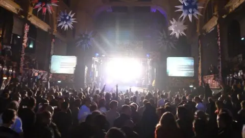 Jon Bryant A large crowded hall with people standing in front of a stage. There are screens on either side of the stage.