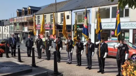 Eight men stand in a line on a town street in suits, holding a large flag each.