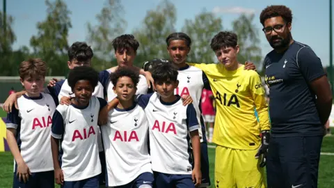 Eight boys in Spurs football kit stand arm in arm in two rows beside their coach. They are on a football field. All of them are looking into the camera. 