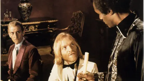 Getty Images Tom Chadbon sits in the middle dressed in a dinner jacket. He has long blonde hair and beard. Calvin Lockhart is stood talking to him while holding a candle. Peter Cushing sits to his right.