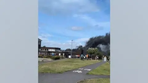 Shanklea Primary School during the fire. The building is two storeys tall and made of dark brick. Large plumes of black smoke can be seen billowing out of the building.