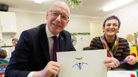 Getty Images John Swinney on a school visit holding a fairly crude picture of himself that you rather hope was drawn by a child