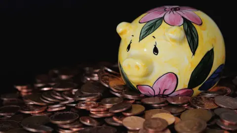 PA Media A yellow money bank pig painted with pink flowers and green leaves sat on a table covered in brown coins. The background is black.