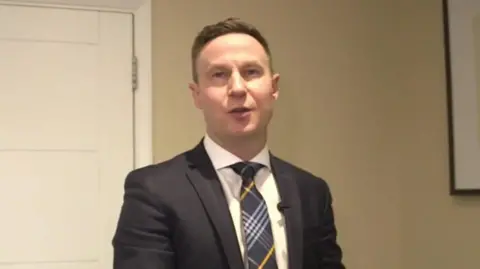 Matthew Uden is seen in a suit standing in his funeral parlour in front of a plain wall and white door.