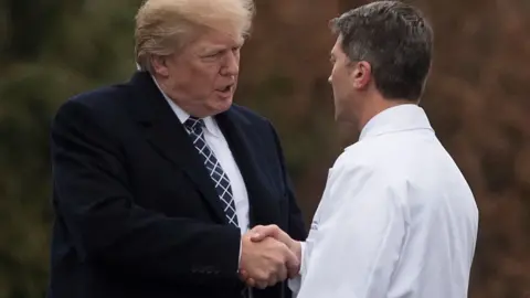 Donald Trump in a suit shaking hands with a man in a white coat