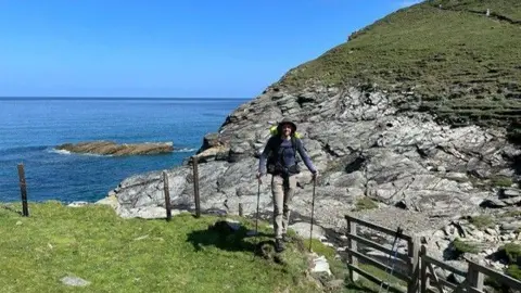 Handout Aidan Courts walking on a footpath by a cliff and the sea. there are fenceposts and a wooden gate near him. The sea is blue in the background. His is wearing a hat and walking gear.