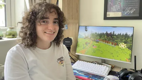 Seren Nia Edwards A brown curly haired girl smiling down the barrel of the camera. Behind her, is a computer monitor showing a replicated version of the Wrexham Eisteddfod in a virtual Minecraft world.