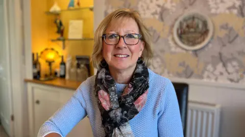 A woman sits in the dining room of a guesthouse. It has colourful wallpaper and bookcase in the background that is lit. 