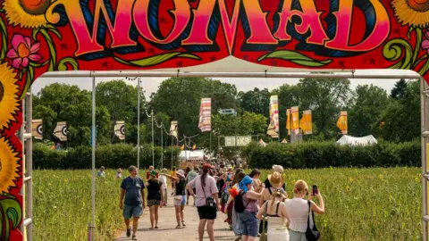 Garry Jones Photography A crowd of people walk under a large pink sign with the word 'WOMAD' and large sunflowers painted on it. They are walking down a long path through a field of sunflowers, strung with lights and the festival site visible in the background. 