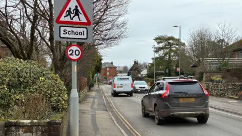A residential road. There are vehicles on either side of the road. Two signs -one saying 20mph and the other saying 'school' are attached to a post. There are buildings and stone walls either side of the road.