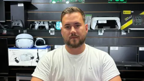 There is a man with a white t-shirt, dark brown hair, a short beard and a straight face. Behind him is technolgy which includes a black xbox, playstation controllers, a teal Nintendo switch, a printer, a Playstation VR and multiple laptops. All of this is in front of a black wall.
