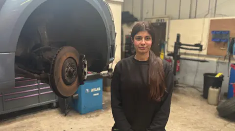 Sophie stops in front of a stripped wheel arc. She has a long brown hair on her shoulder in a ponytail. She uses a black sweater.