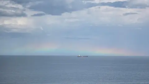 Fish Eyes The perfectly straight horizon of the deep-blue sea at Hartlepool. In the distance is a ship. Clouds hang above. However, just above the water, there appears to be floating a rainbow-coloured haze - rain is reflecting in the distance.