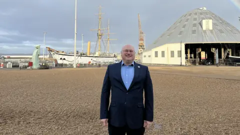 A man in a suit stands on a pebble beach. He is smiling widely. 