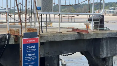 A close up photo of the landing stage at Gorey pier. A closed sign is visible on it. Long beach can be seen behind it.