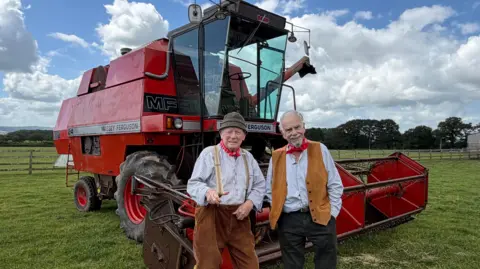 Two elderly men wearing red neckerchiefs and white shirts, standing in front of a red combine harvester in a field