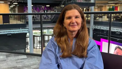Johanna Robinson looks at the camera. She has long brown hair and is wearing a blue shirt, and is sat on a sofa in BBC central square where you can see the office and BBC screens in the background. The picture is cropped so you can see her face and shoulders