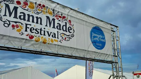 A large steel frame with a white banner with the Mann Made festival logo on it,  which has black writing and yellow and red drawings of foodstuffs such oranges and apples surrounding it.