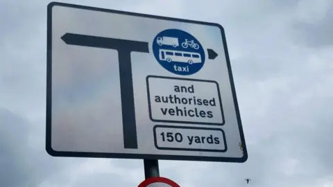 Bus gate sign in Aberdeen, depicting road junction and sign showing lorries, bikes, buses and taxis allowed, with a seagull flying in the background.