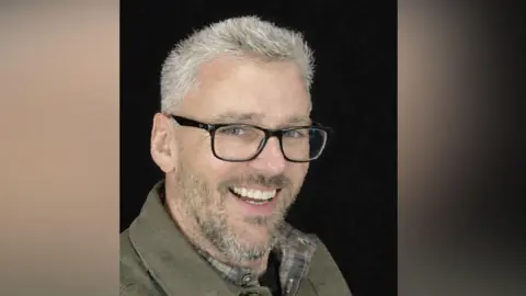 Craig Hurcombe smiles at the camera. He has short grey hair and is wearing black, thick-rimmed glasses and a brown shirt. He is pictured against a black background.