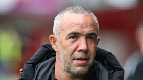 SNS Dougie Imrie, a man with grey/silver hair and beard, standing on the touchline at a football game