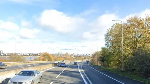 Cars drive down a three-lane motorway on a sunny day with green trees on the side. The Leeds city skyline is in the distance.