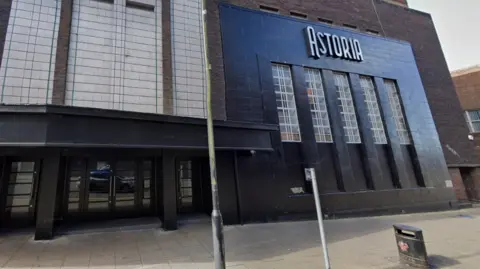 The venue has a blue brick and glass entrance on a main street in the city, with a large sign that reads Astoria.