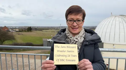 BBC East Midlands weather presenter Sara Blizzard, holding a gold plaque that reads 'The Sara Blizzard Weather Station, Celebrating 26 Years of BBC Forecasts' 