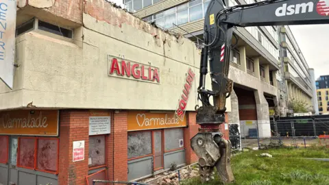 A yellow and black digger on caterpillar tracks is positioned in front of a concrete and glass low-rise building, with has ANGLIA SQUARE in red letters, with a shop underneath it. The SQUARE SECTION has partially detached and is hanging loose from the wall. There are taller buildings to the right of the heavy plant.