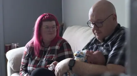 Tim Brentnall wearing a checked black and white shirt, bald and wearing glasses. He is looking down at baby Lando who he is holding in one arm, with a dummy in the other hand. Sitting on the same sofa next to him is his partner Steph who is also in a checked shirt but black white and red, she has pink dyed hair and is looking lovingly at baby Lando