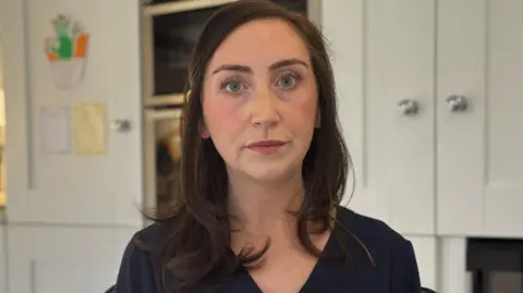Niamh is looking into the camera. She has long brown hair and is wearing a navy coloured top. She's standing in a kitchen with cream cabinets behind her.