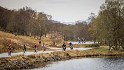 Paul Campbell Cyclists negotiate a twisting road along the shores of a Highland loch. The surrounding landscape is brown moorland, silver birch woodland and in the background is a mountain with snow on it.