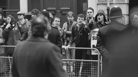 Getty Images A black and white image take during the miners' strike. There are a row of angry looking men stood behind a metal fence. Two of the men are pointing their fingers at a man stood in front of them. There is a policeman stood at the side with his arms behind his back. 