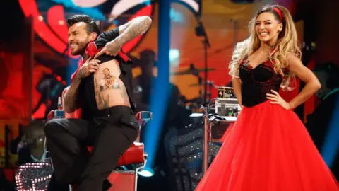 Pete Wicks with medium-length swept-back black hair, wearing a black shirt, red scarf and black trousers, performing a twisting manoeuvre with Jowita Przystal with long blonde hair wearing a dress with black top and red skirt. The Strictly band is visible behind them with art deco shapes on the scenery.