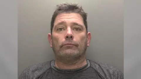A police custody picture showing the head and shoulders of a man with short brown hair and stubble wearing a grey T-shirt against a grey backdrop. 