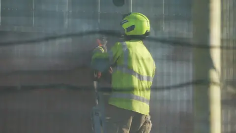 Jamie Niblock/BBC The back view of a firefighter in a hi-vis yellow jacket, standing on the top of a ladder at Highpoint Prison. The shot is taken through a wire fence. 