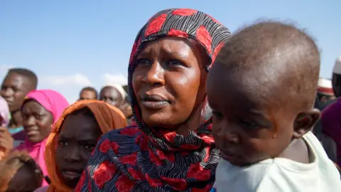 Joyce Liu / BBC A woman in a red and black flowery headscarf carries her baby with several people standing around her