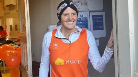 A woman stood in a doorway and smiling wearing an orange top that says 'Maggie's'