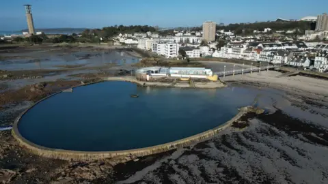 The Havre des Pas lido during low tide on a sunny day. The pool is circle-shaped and is filled with water. St Helier is in the background.