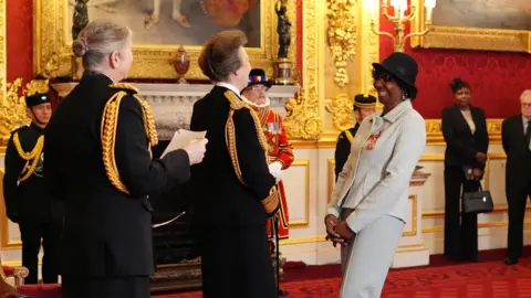 PA Media Princess Anne faces Deta Hedman. Anne is in her ceremonial black uniform and Deta is in a white or light-coloured blazer and skirt. She has a black hat on. They are in an ornate hall, with red walls and gold picture frames on the back wall.