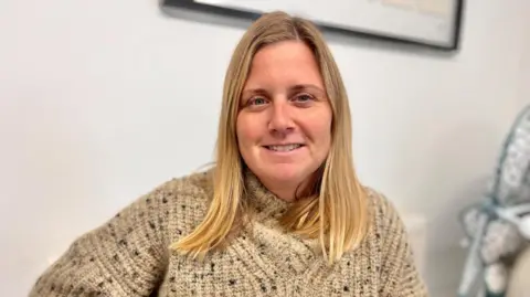 Martin Giles/BBC Emma Spencer is sitting at a table at the Lakenheath Community Center wearing a thick, beige knitted sweater with a speckled pattern. In the background, part of a framed picture is visible on a white wall. Emma is smiling at the camera and look relaxed.