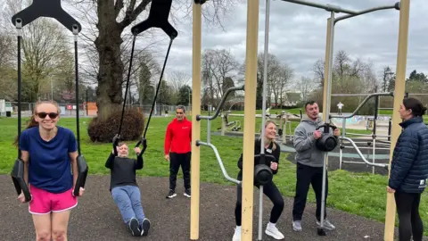 A group using outdoor gym lit under an instructor's direction