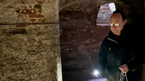 A man stands in a dark basement and he is wearing trousers, an anorak and holding a torch and some keys, as he studies an old stone wall in front of him