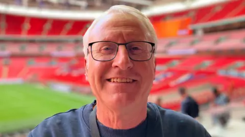 Lewis Adams/BBC Glenn Pennyfather wearing glasses with black rims and a navy sweater. He is standing in front of a blurred Wembley Stadium pitch.