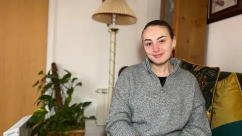 Antonia sits on a sofa and smiles at the camera. She is wearing a grey zip-up fleece with a black top underneath and a necklace. There is a small plant and tall lamp to her left and a cabinet behind her. She has dark brown hair which is tied back.