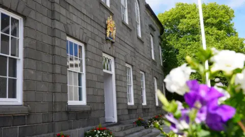 A grey bricked building with sash windows and a coat of arms of the wall. A set of purple and white flowers are in the foreground.