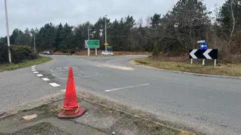 A large roundabout with trees in the centre. In the foreground is a red traffic cone. There is a patch of sand spread across the right hand lane of the roundabout.