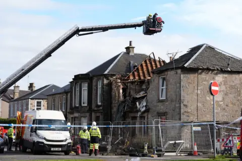 PA Media Firefighters connected  a crane survey   the remains of a level  destroyed successful  an explosion. The foreground is cordoned disconnected  and exigency  work  workers are seen beside the constabulary  cordon. 