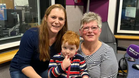 Isaac Chenery/BBC A head and shoulders image of three people pictured in a radio studio. A woman with grey short hair and glasses, and wearing a dark grey and white striped top, is seated, with a three-year-old boy on her lap. He has red hair and is wearing a blue, red, black and and white striped fleece. The tip of his right index finger is in his mouth. Crouching to his level, to his right, is a woman with long red hair, dressed all in blue. All three are looking at the camera. 