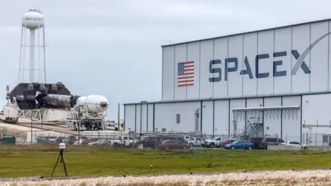 A view of a warehouse with the US flag and the word 'SpaceX' on the side with the Falcon 9 rocket in the background against a cloudy sky at the Kennedy Space Center in Cape Canaveral, Florida, as it is moved a day before launch on 14 January 2026.